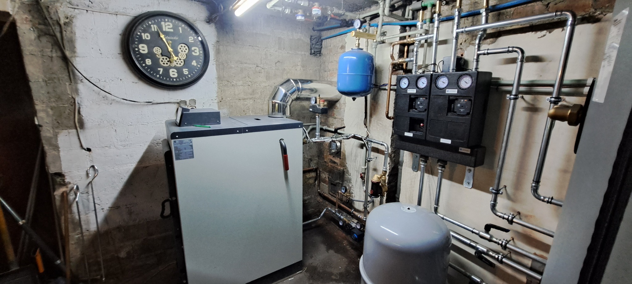 Kotłownia z kotłem grzewczym, zegarem ściennym i rozbudowaną instalacją hydrauliczną z rur, zaworów i manometrów w piwnicy o surowych ścianach.