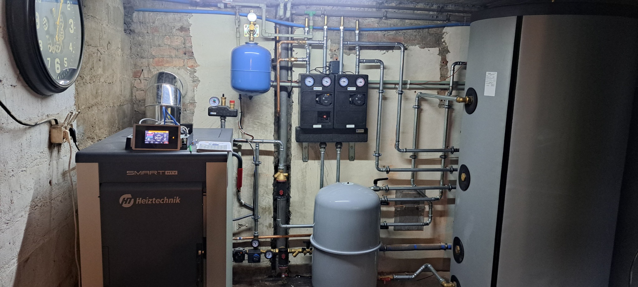 Kotłownia z piecem na pellet Heiztechnik Smart HTV, zbiornikiem akumulacyjnym, naczyniami przeponowymi i rozbudowaną instalacją hydrauliczną z manometrami na tle ściany z widoczną cegłą.