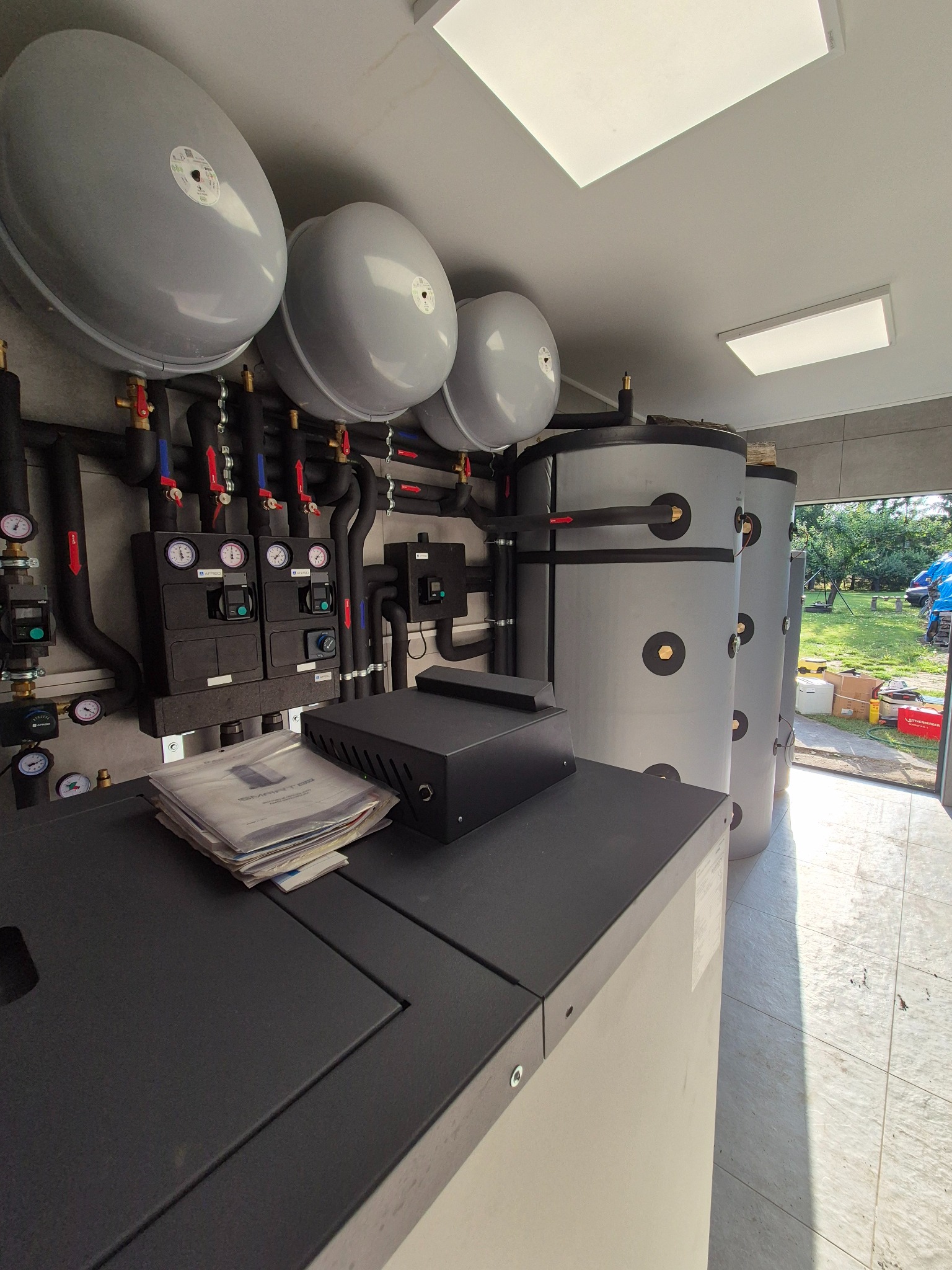Wnętrze kotłowni z trzema zbiornikami akumulacyjnymi, czarnymi rurami, manometrami i automatyką. Na pierwszym planie dokumentacja techniczna i czarna obudowa sterownika.