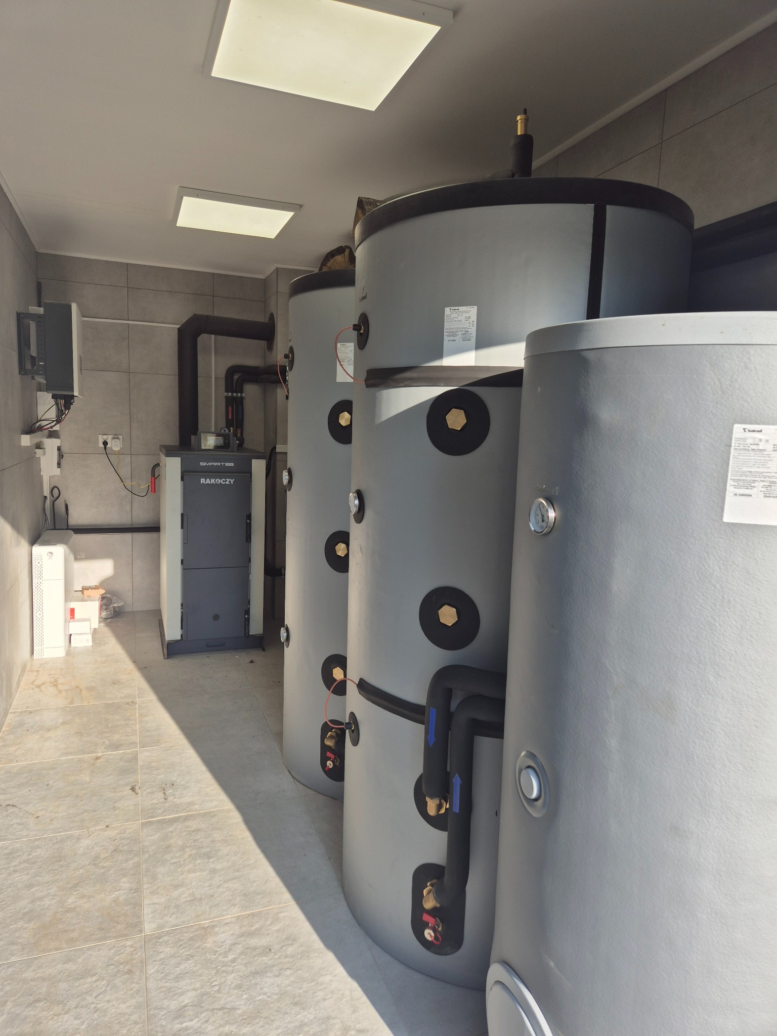 Kotłownia z piecem RAKOCZY i trzema szarymi zbiornikami akumulacyjnymi, połączonymi czarnymi rurami. Na ścianie widoczny panel sterowania. Pomieszczenie wyłożone jasnymi płytkami.