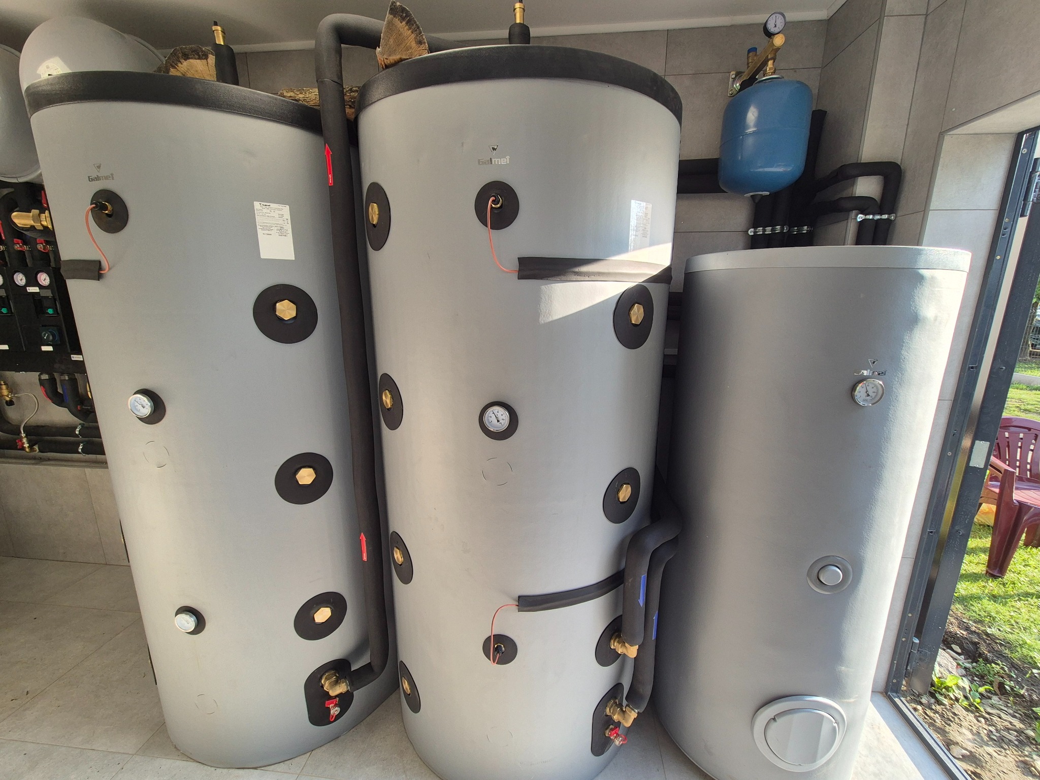 Szereg szarych, cylindrycznych zbiorników Galmet w kotłowni. Widoczne czarne rury, manometry i zawory. Na zbiorniku leżą kawałki drewna. Na zewnątrz trawa i krzesło.