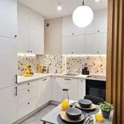 Nowoczesna kuchnia z białymi szafkami, blatem w stylu lastryko i drewnianymi lamelami oddzielającymi przestrzeń jadalną, nakryty stół z sokiem pomarańczowym i owocami na blacie.