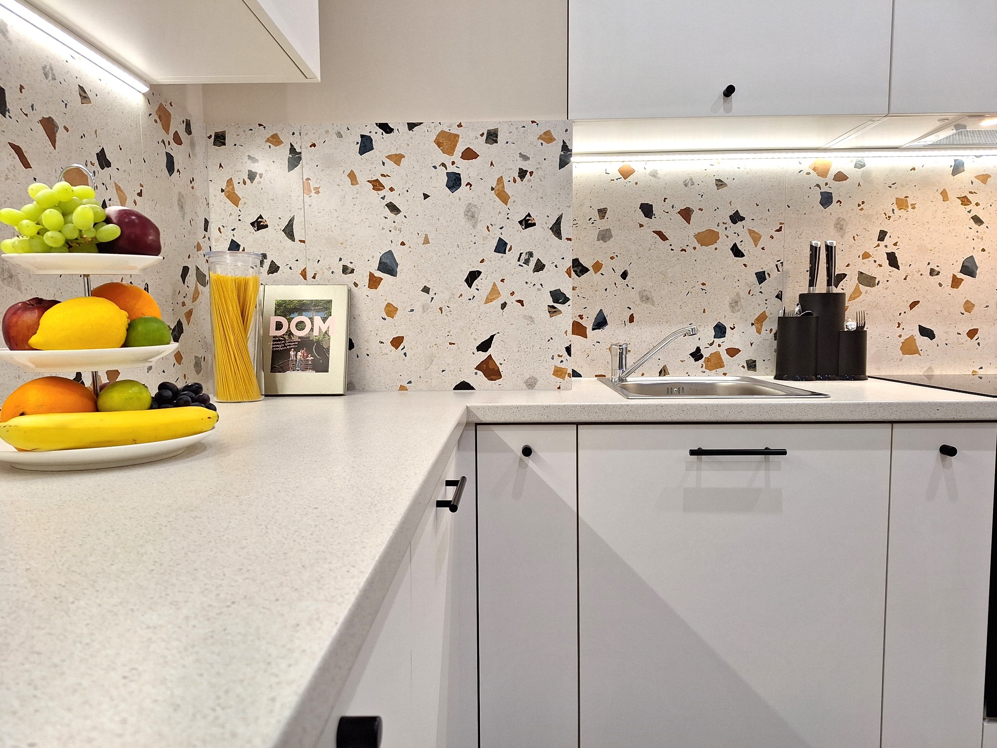 Nowoczesna, jasna kuchnia z białymi szafkami, blatem w drobny wzór i dekoracyjnym panelem ściennym w stylu lastryko. Na blacie misa z owocami, słoik z makaronem i stojak na noże.