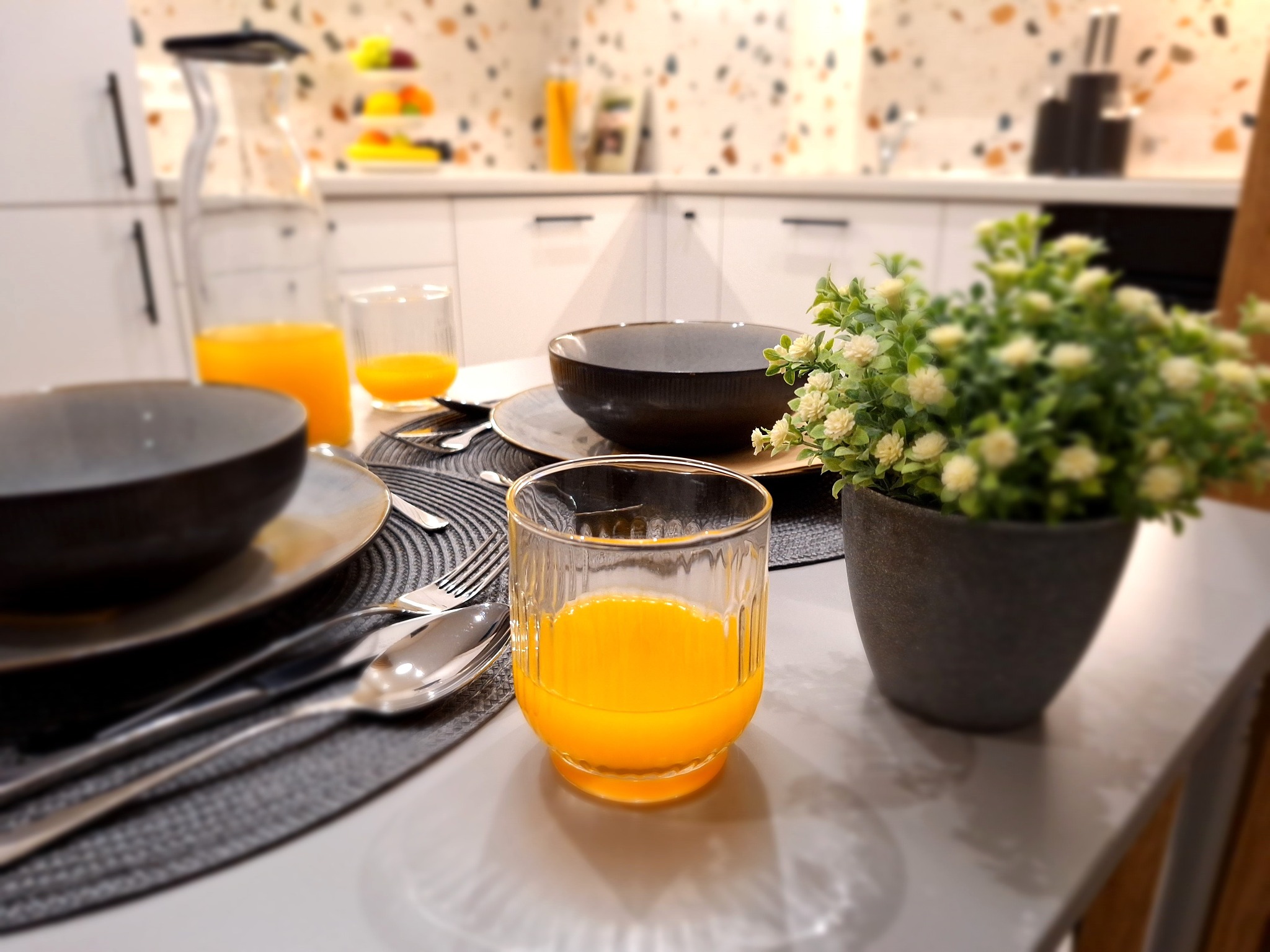 Nowoczesna kuchnia z blatem śniadaniowym, zastawą stołową, szklankami z pomarańczowym sokiem i doniczką z białymi kwiatami na pierwszym planie, w tle białe szafki z czarnymi uchwytami.