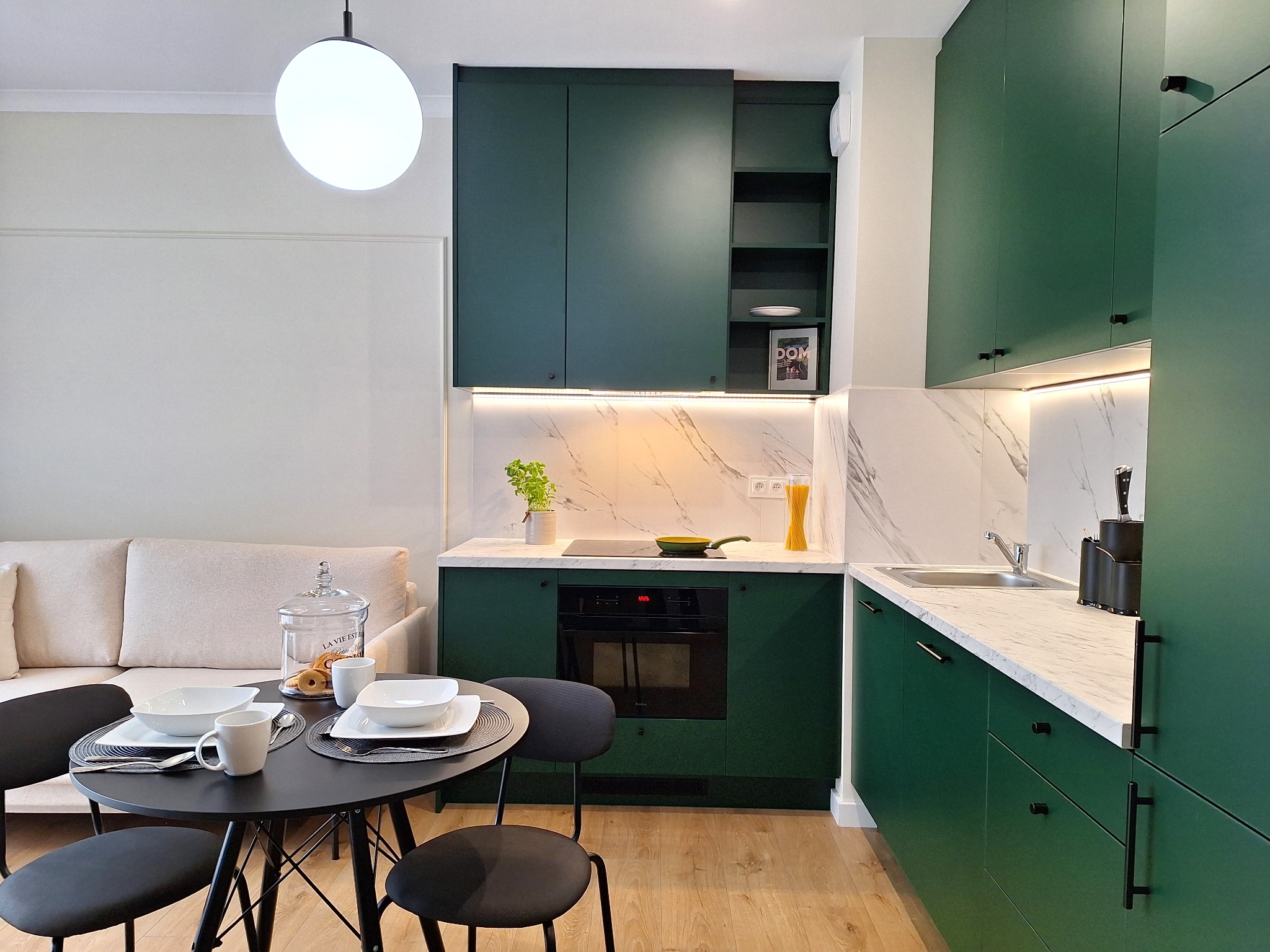 Nowoczesna kuchnia w odcieniach butelkowej zieleni z marmurowym blatem, okrągłym stołem jadalnym i minimalistycznym oświetleniem, ukazująca aranżację wnętrza gotową do zamieszkania.