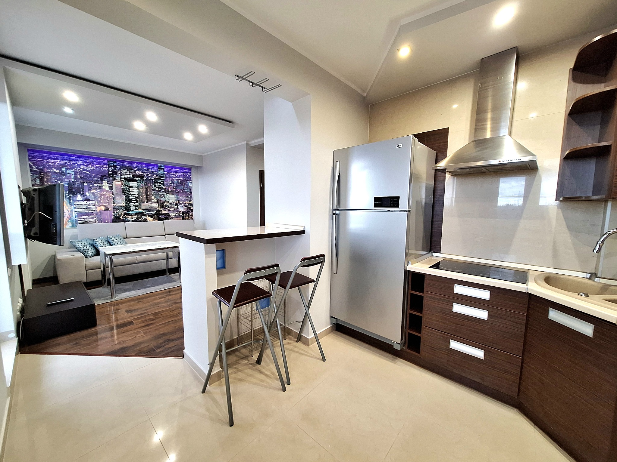 Nowoczesna kuchnia z aneksem w Gorzowie Wielkopolskim. Widoczna zabudowa kuchenna z ciemnego drewna, srebrna lodówka, okap, dwie składane hokery przy blacie oddzielającym kuchnię od salonu z sofą...