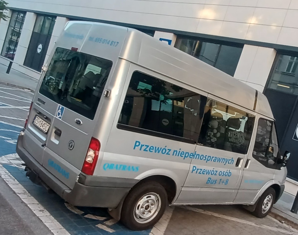 Srebrny Ford Transit przystosowany do przewozu osób niepełnosprawnych, zaparkowany na niebieskim miejscu parkingowym dla osób z niepełnosprawnościami, z napisem 'Przewóz osób. Bus 1+8' na boku.