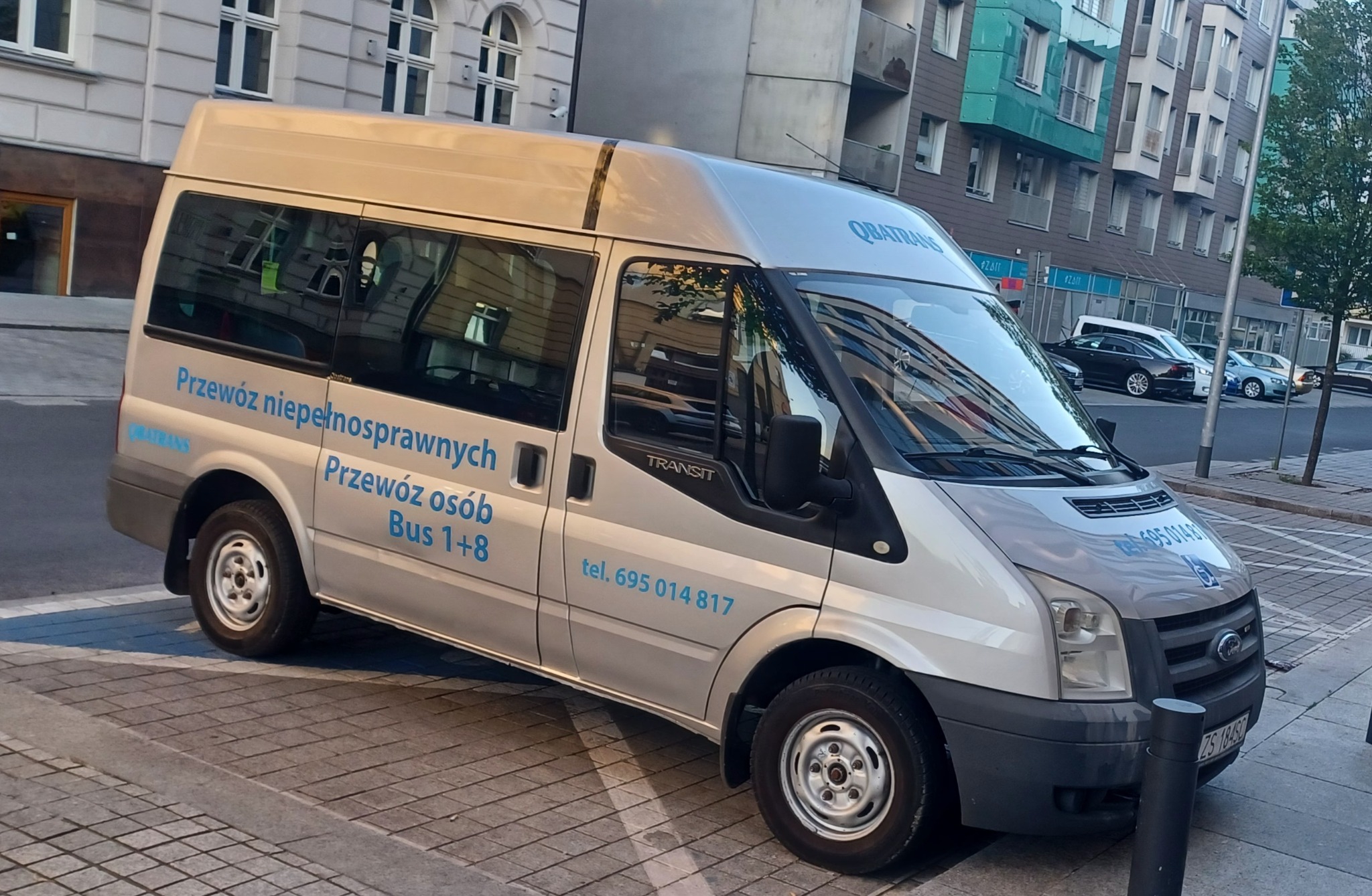 Srebrny bus Ford Transit z niebieskimi napisami 'Przewóz niepełnosprawnych', 'Przewóz osób Bus 1+8' oraz numerem telefonu, zaparkowany na chodniku z kostki brukowej na tle budynku miejskiego.