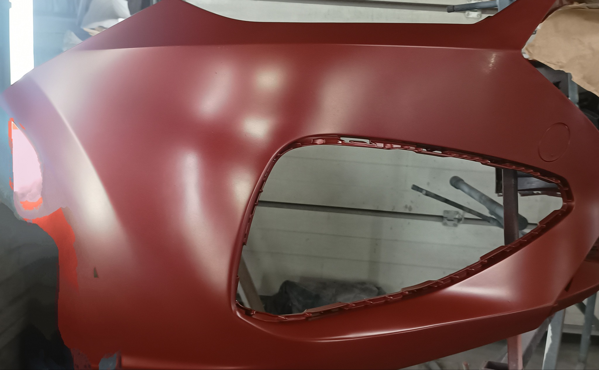 Czerwony, świeżo polakierowany zderzak samochodowy na stojaku w warsztacie, widoczne ślady szpachli w lewym rogu.