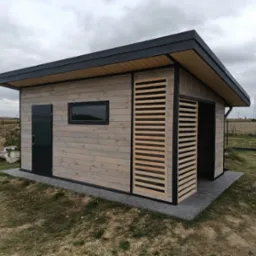 Nowoczesna, drewniana altana z elementami żaluzjowymi w Rusiłach, z czarnymi detalami i płaskim dachem, otoczona trawnikiem i kostką brukową.