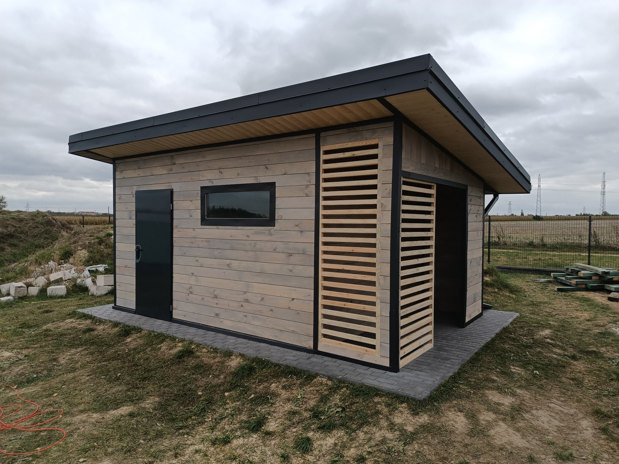 Nowoczesna, drewniana altana z elementami żaluzjowymi w Rusiłach, z czarnymi detalami i płaskim dachem, otoczona trawnikiem i kostką brukową.