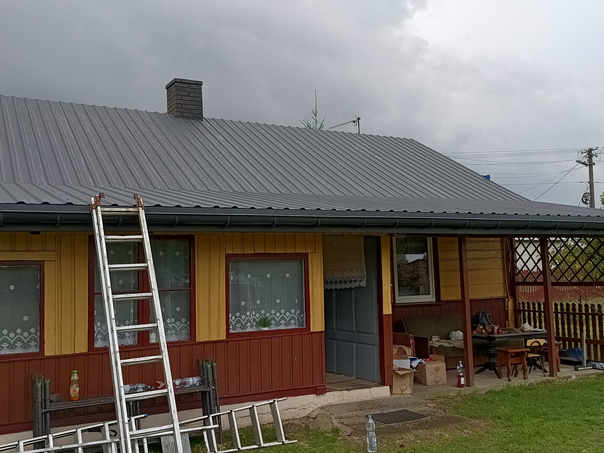 Dom z nowym, szarym dachem z blachy trapezowej, na tle pochmurnego nieba. Przystawiona drabina aluminiowa, częściowo rozłożona, z butelkami na dolnych szczeblach. Widoczny komin z cegły i antena...