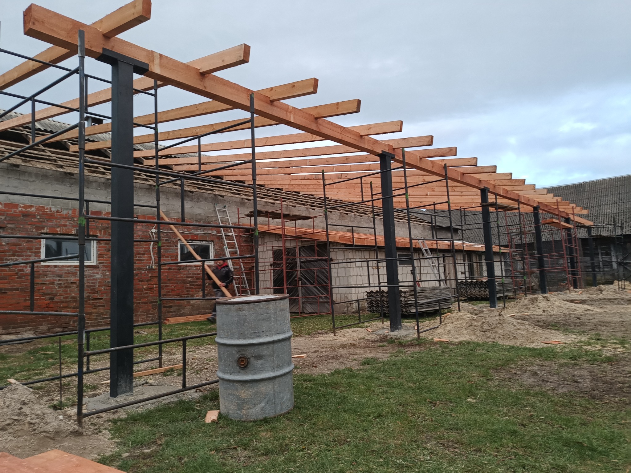 Montaż drewnianej pergoli z elementami metalowymi, widoczne czarne słupy i drewniane belki na tle ceglanego budynku i pochmurnego nieba, w tle drabina i osoba niosąca drewno.