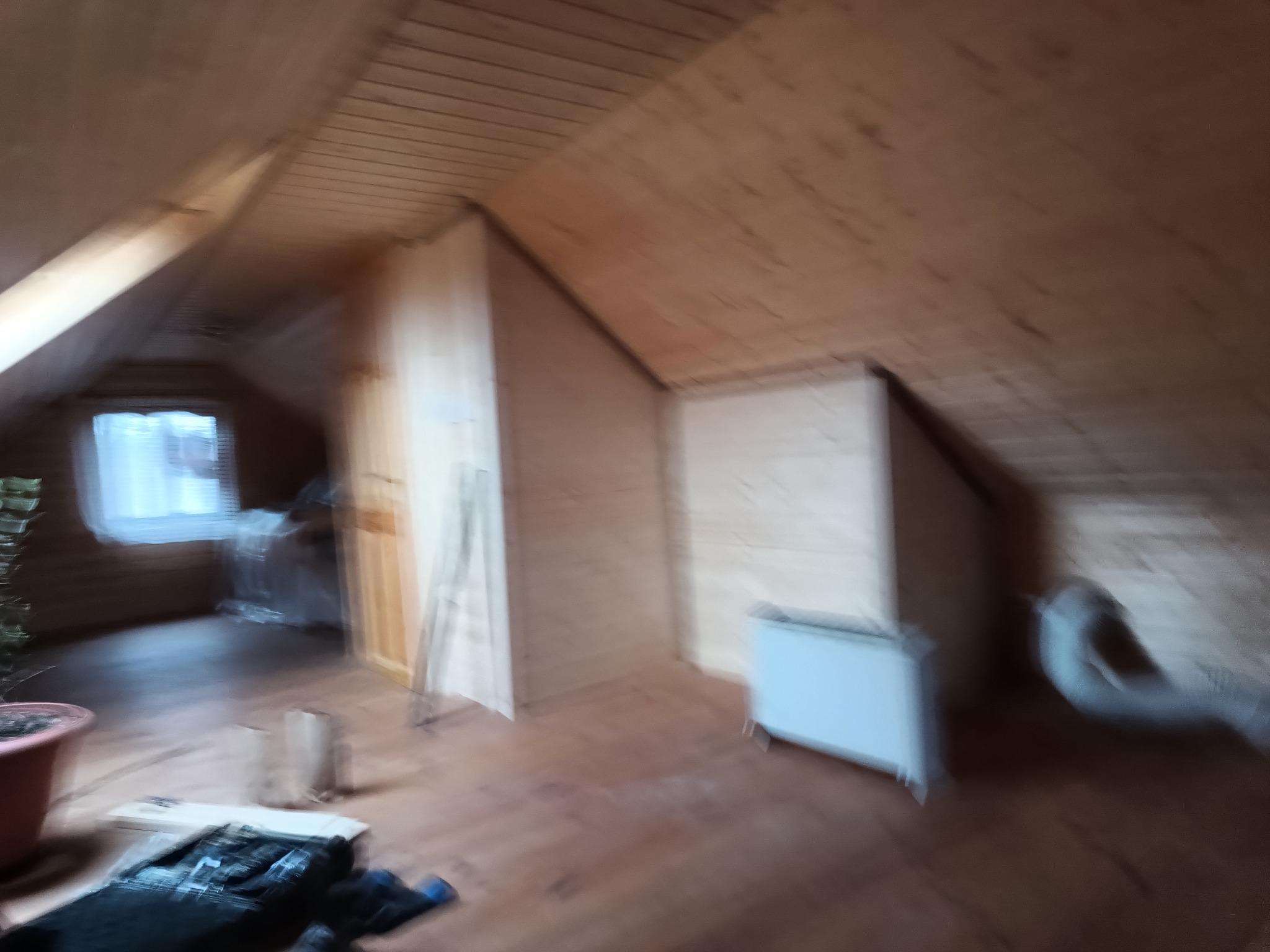 Poddasze z drewnianym wykończeniem, widoczne okno, grzejnik i częściowo zasłonięte przedmioty, w trakcie prac wykończeniowych.