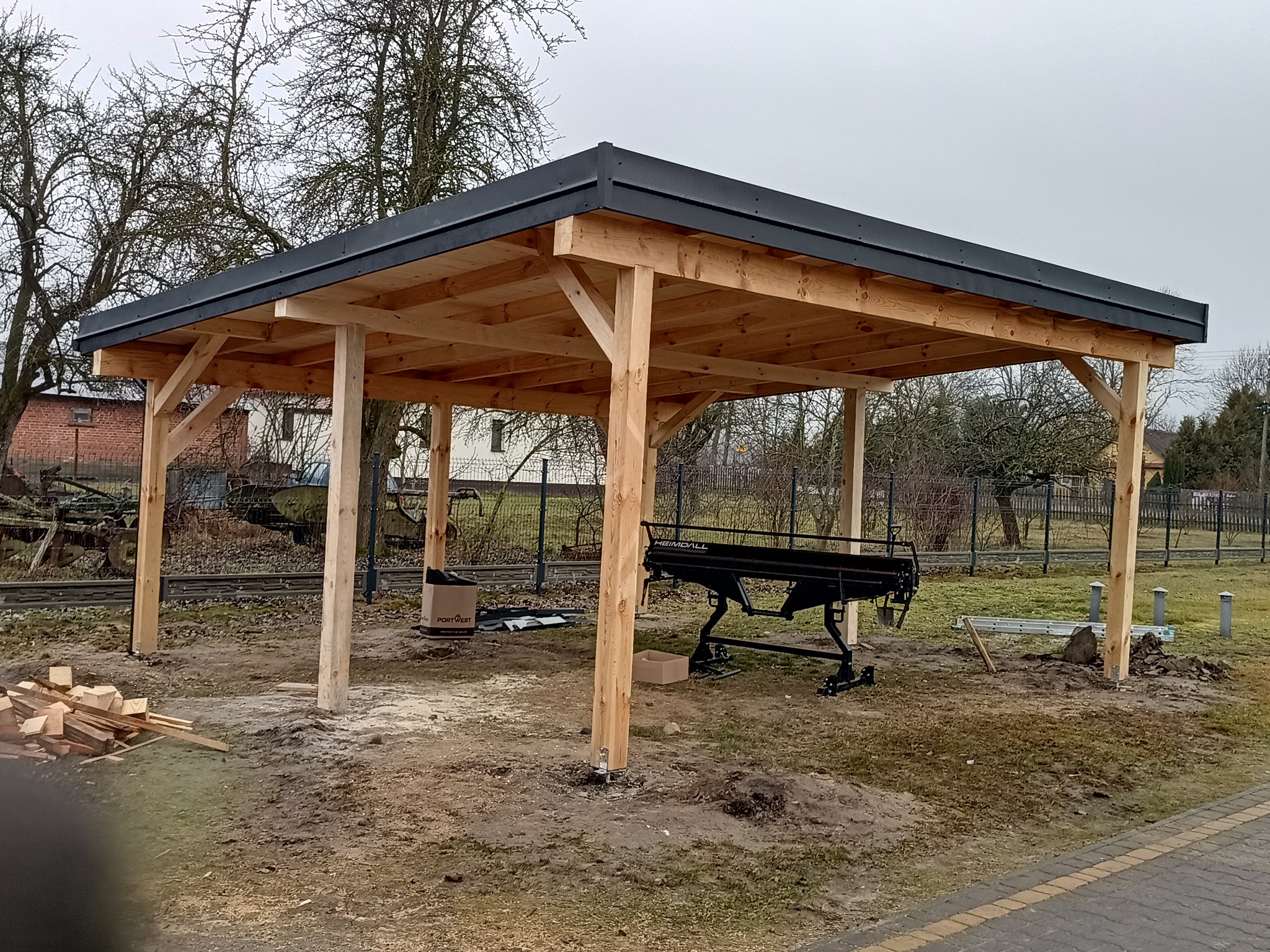 Drewniana konstrukcja z czarnym dachem w trakcie budowy, widoczne narzędzia i materiały budowlane na ziemi.