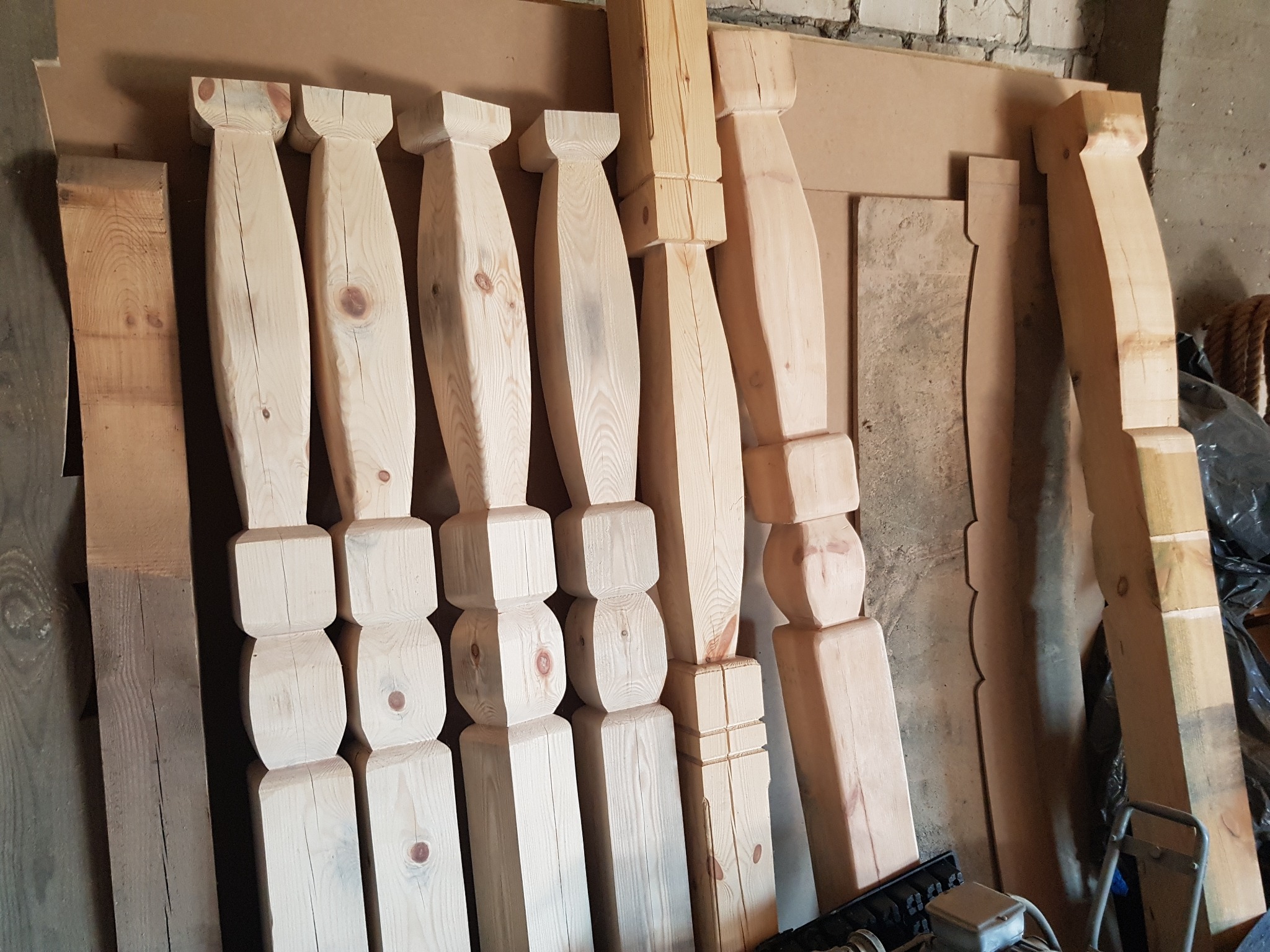 Surowe, nieobrobione drewniane elementy konstrukcyjne o różnych kształtach, oparte o ścianę z cegieł, czekające na montaż lub obróbkę.