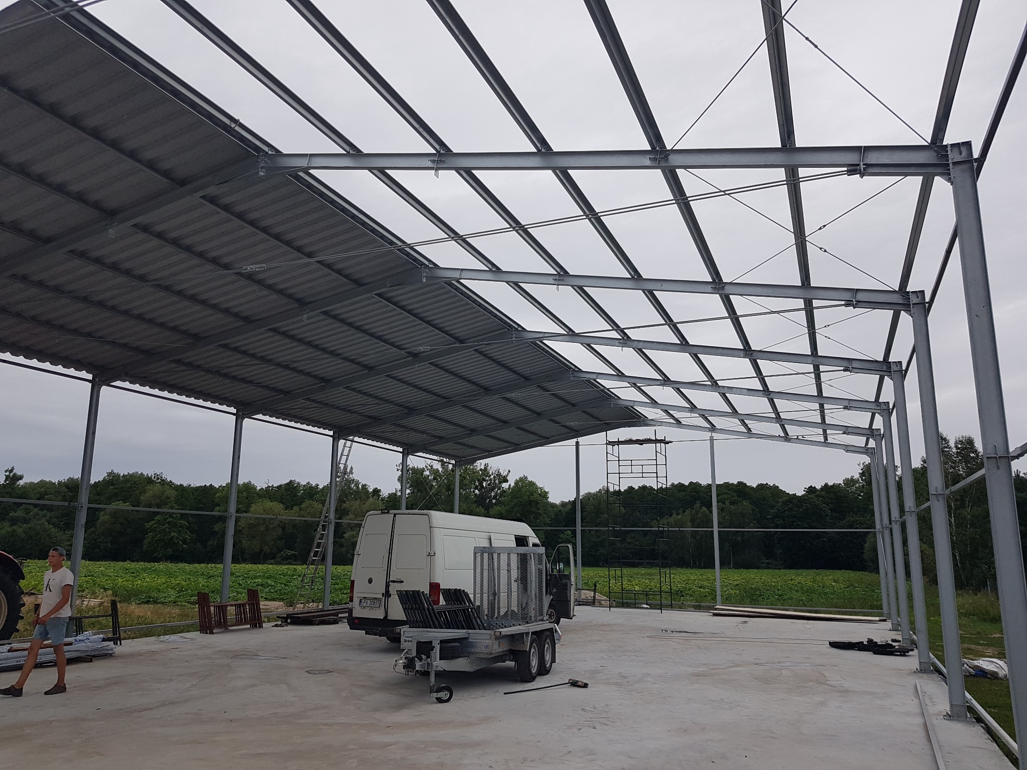 Konstrukcja stalowa z częściowo zamontowanym dachem z blachy falistej, widoczny biały samochód dostawczy i przyczepka z elementami konstrukcyjnymi, w tle pole i las.