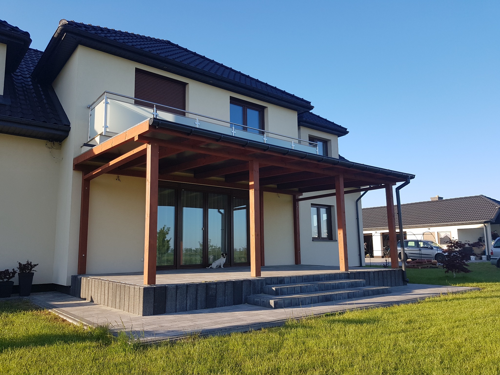 Nowoczesny dom z drewnianą pergolą zadaszającą taras, z psem siedzącym na tarasie, widok z trawnika w słoneczny dzień.