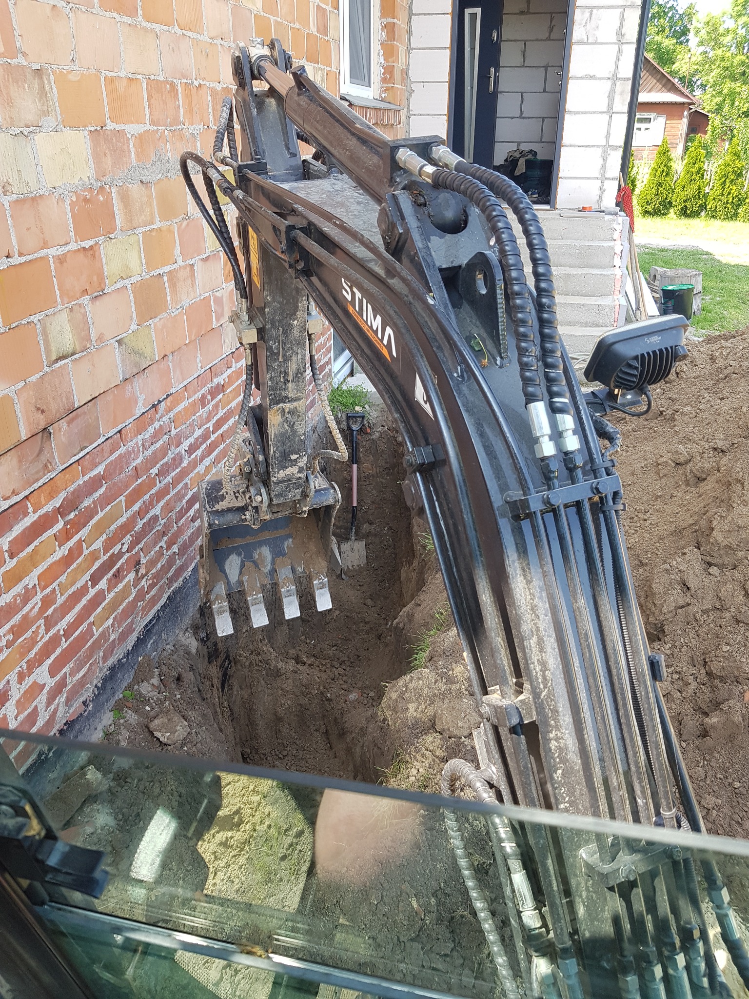 Mini koparka marki Stima wykonuje wykop wąskoprzestrzenny wzdłuż ceglanej ściany budynku, widok z kabiny operatora, obok stoi łopata.