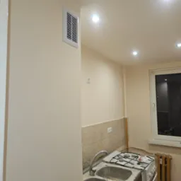 Świeżo pomalowana ściana w kuchni w odcieniu beżu z kratką wentylacyjną, widoczny fragment starej zabudowy kuchennej i okno.