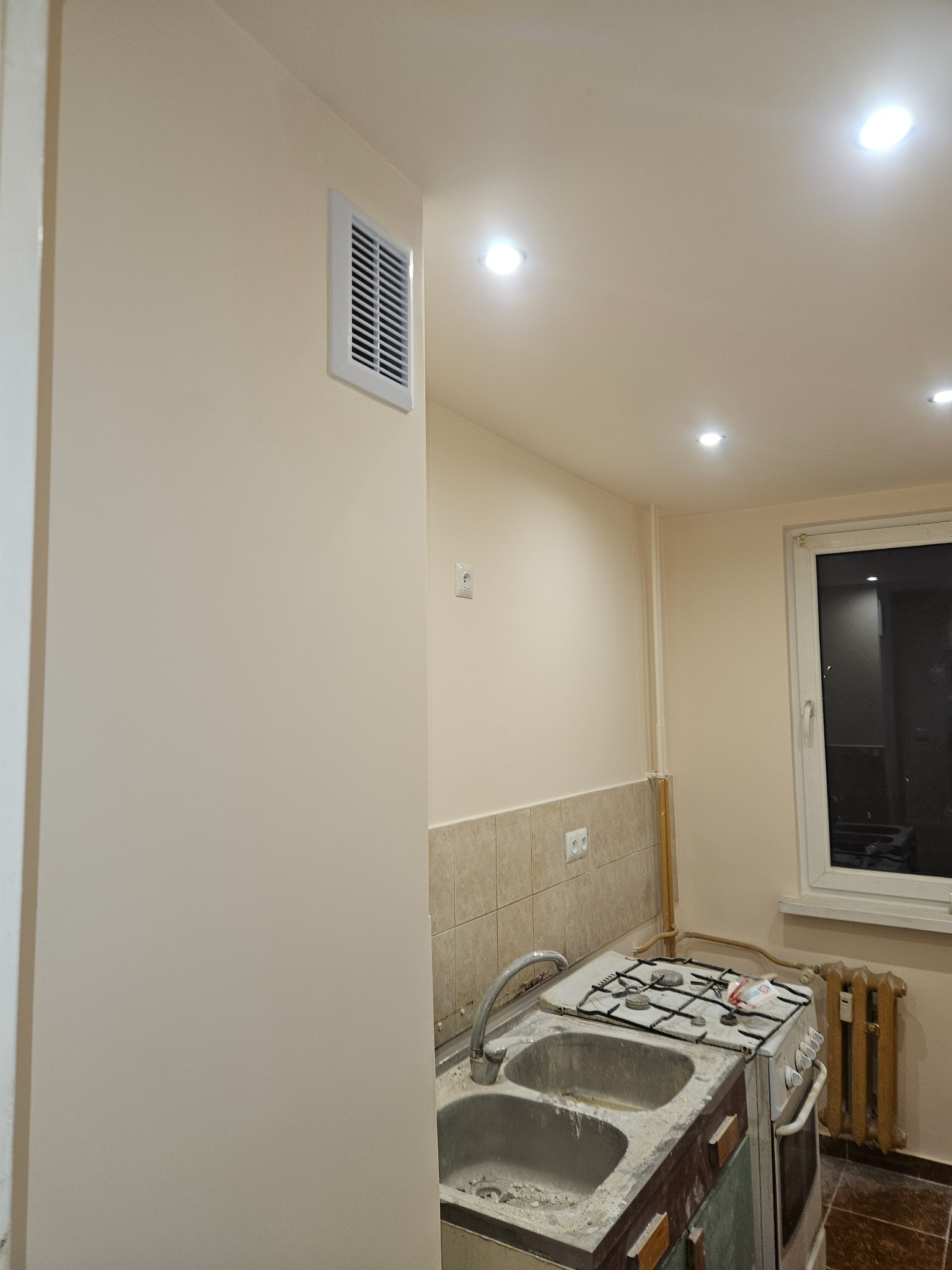 Świeżo pomalowana ściana w kuchni w odcieniu beżu z kratką wentylacyjną, widoczny fragment starej zabudowy kuchennej i okno.