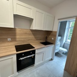 Invest bud - Nowoczesna, jasna kuchnia z drewnianym blatem i czarnym zlewem oraz płytą indukcyjną, widok na częściowo zasłoniętą łazienkę z toaletą w tle.