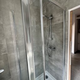 Invest bud - Szklana kabina prysznicowa z chromowanymi profilami i szarymi płytkami w nowoczesnej łazience, widok na biały brodzik i baterię.