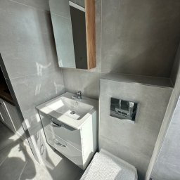 Invest bud - Szara łazienka z białą szafką pod umywalką, lustrem z drewnianym akcentem i toaletą zabezpieczoną folią bąbelkową.