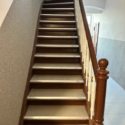 Invest bud - Kręte schody z brązowymi stopniami i białymi balustradami, wykończone aluminiowymi listwami antypoślizgowymi, prowadzące w górę w budynku o ścianach pokrytych szarym tynkiem.