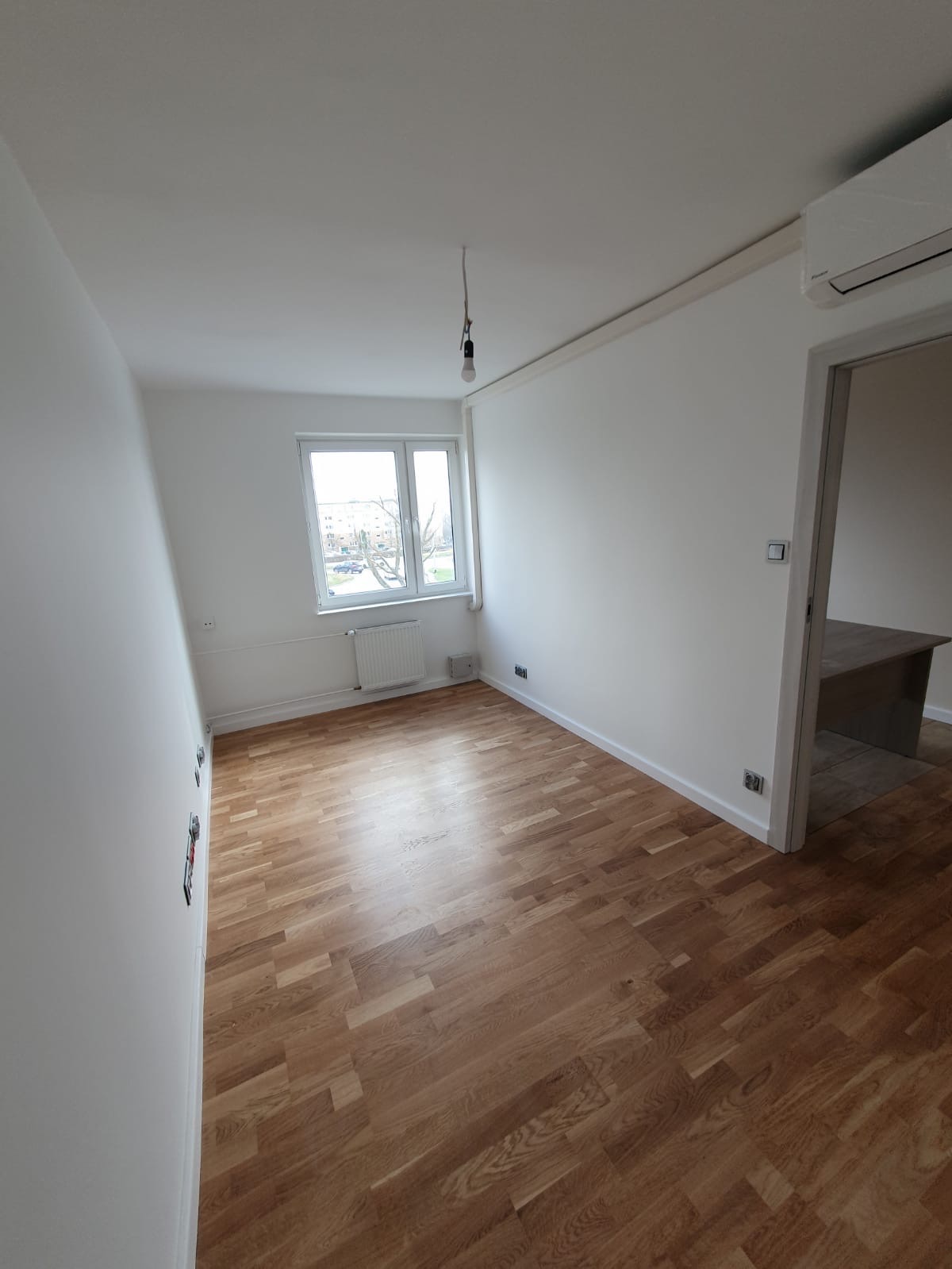 Pusty pokój po remoncie z drewnianą podłogą, białymi ścianami i oknem z widokiem na budynki. Wisząca żarówka bez klosza i widoczny klimatyzator.