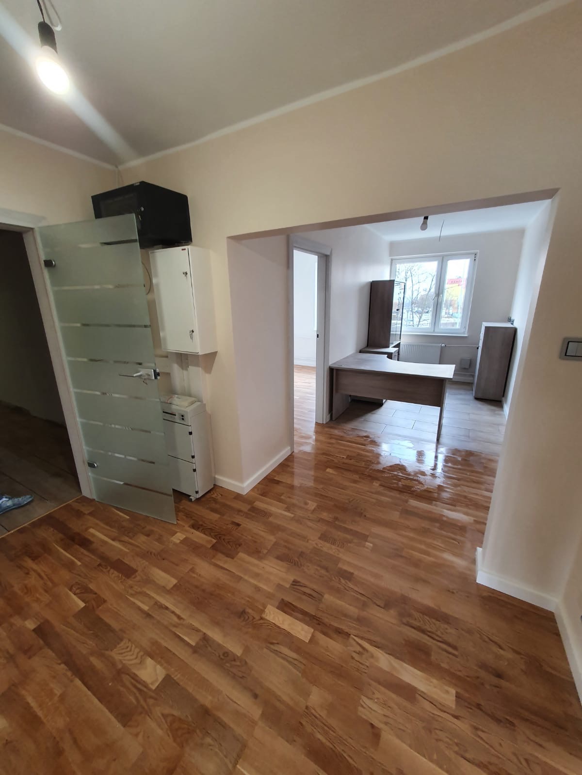 Wnętrze biura po remoncie, widok na odnowioną podłogę z parkietu, świeżo pomalowane ściany w odcieniach beżu i białe listwy przypodłogowe, oraz nowe drzwi z matowego szkła.