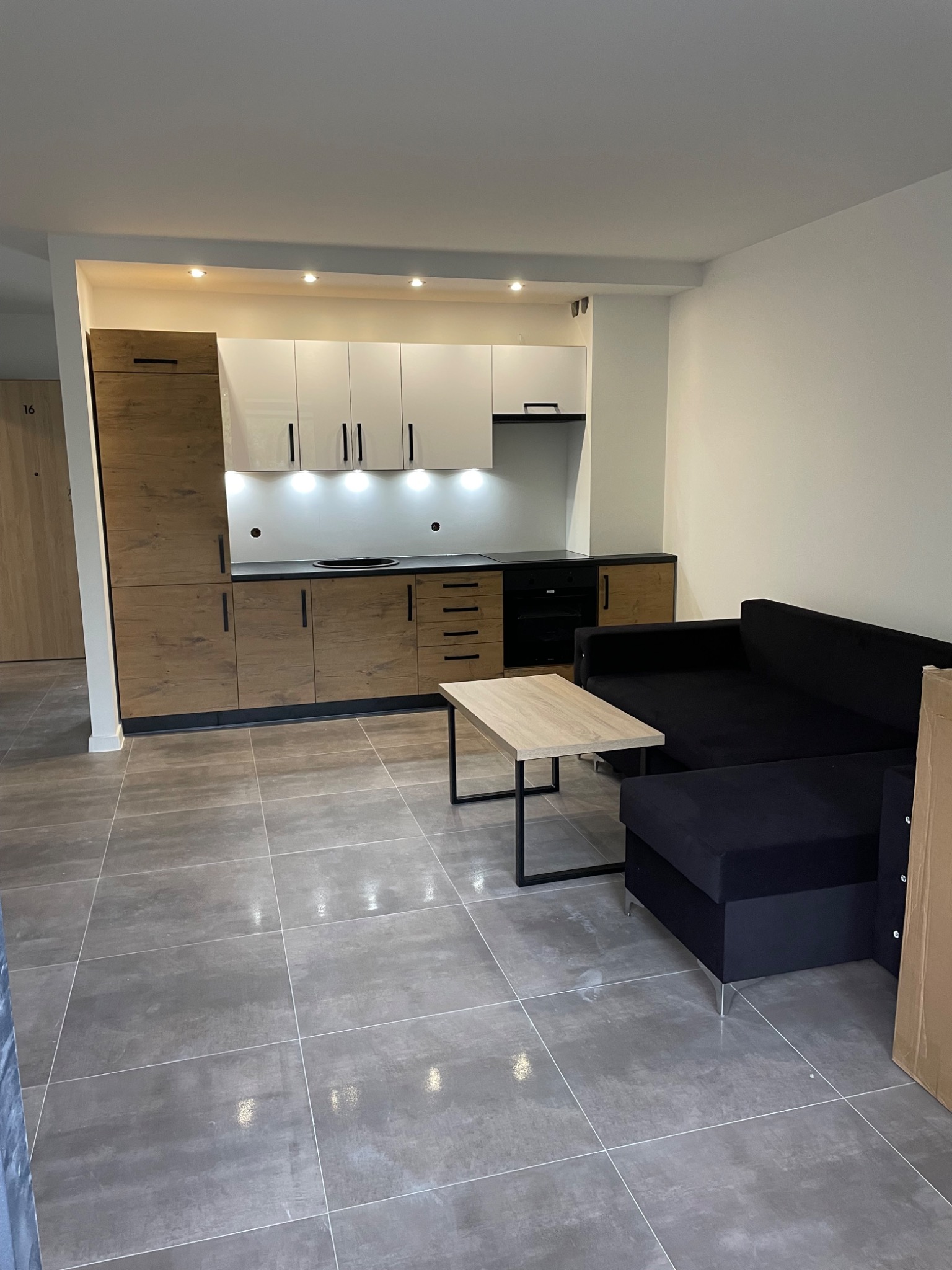 Nowoczesne wnętrze mieszkania z aneksem kuchennym: zabudowa z frontami drewnopodobnymi i białymi, czarna sofa narożna, stolik kawowy z jasnym blatem i szarą podłogą z płytek.