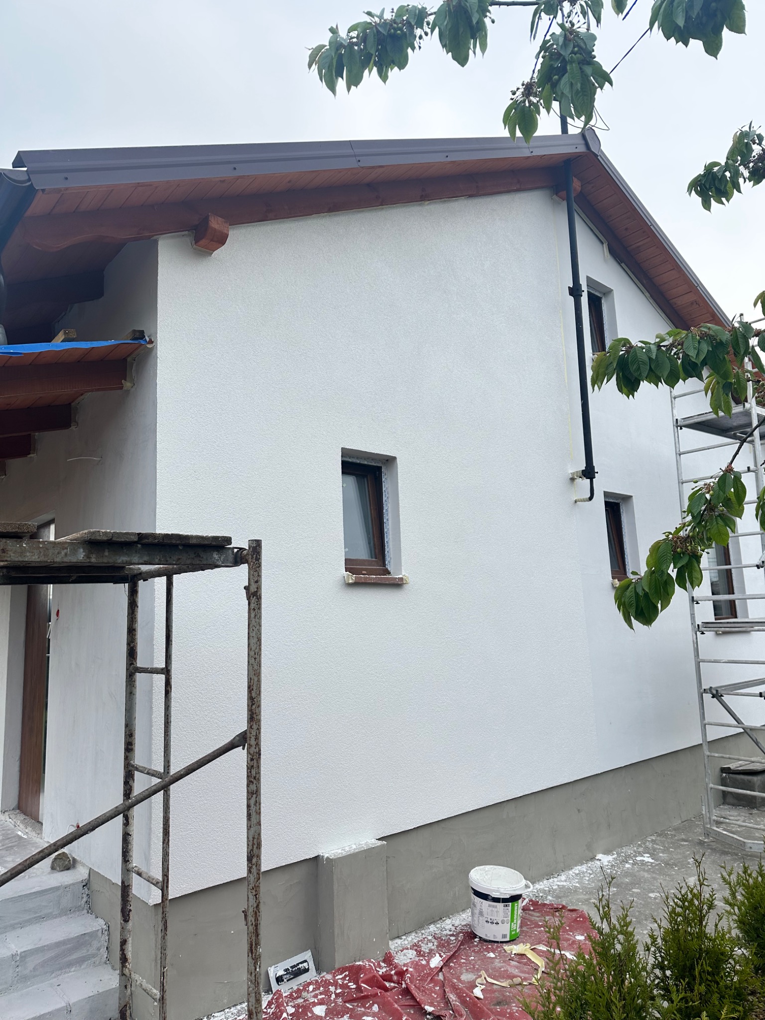Świeżo pomalowana biała elewacja domu jednorodzinnego z brązowymi oknami i dachem, widoczne rusztowania, wiadro z farbą i plandeka zabezpieczająca