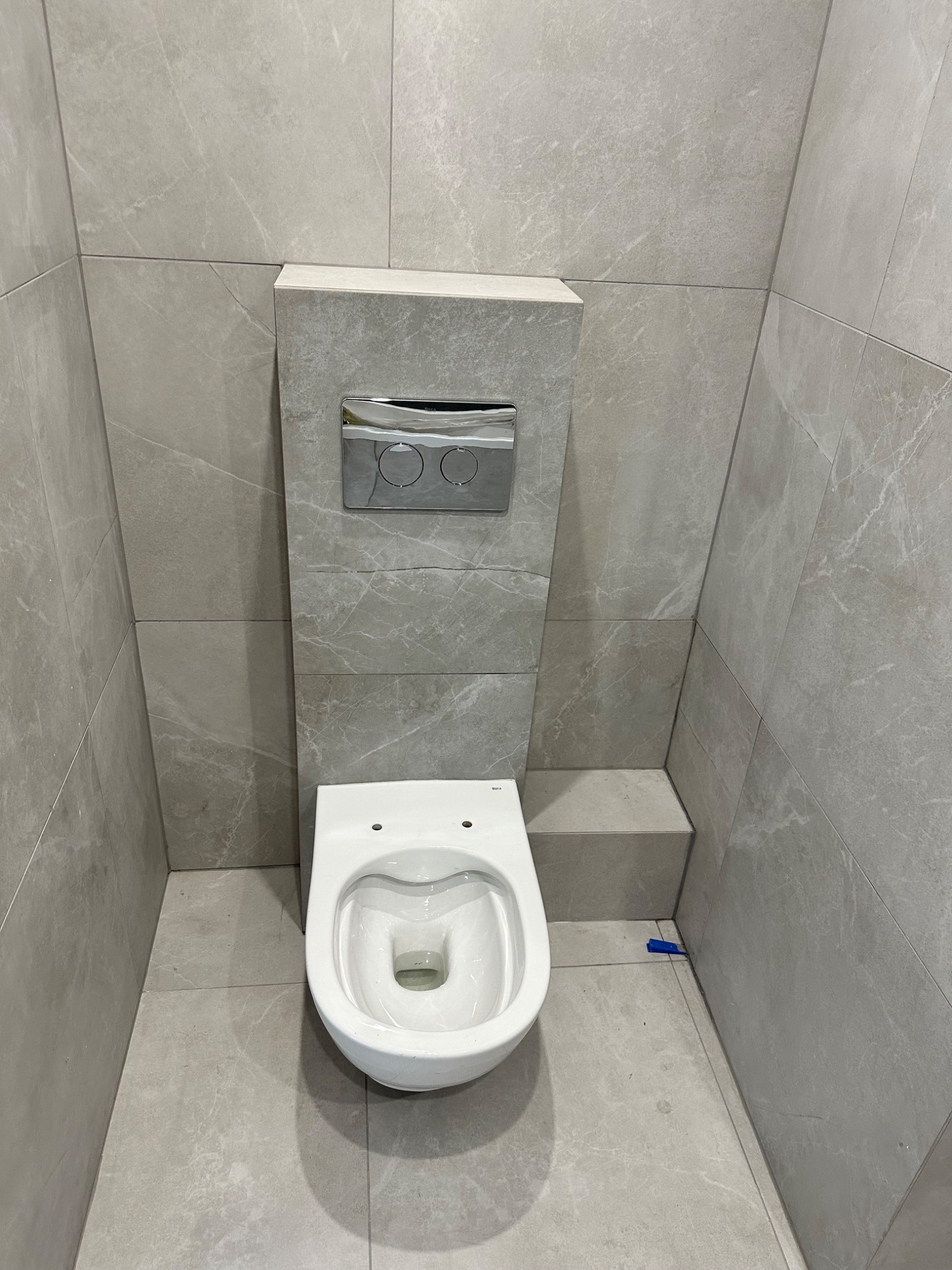 Nowoczesna toaleta podwieszana z chromowanym przyciskiem spłukującym, obudowana płytkami w odcieniach szarości z subtelnym marmurkowym wzorem, widoczna niebieska zaślepka na podłodze.