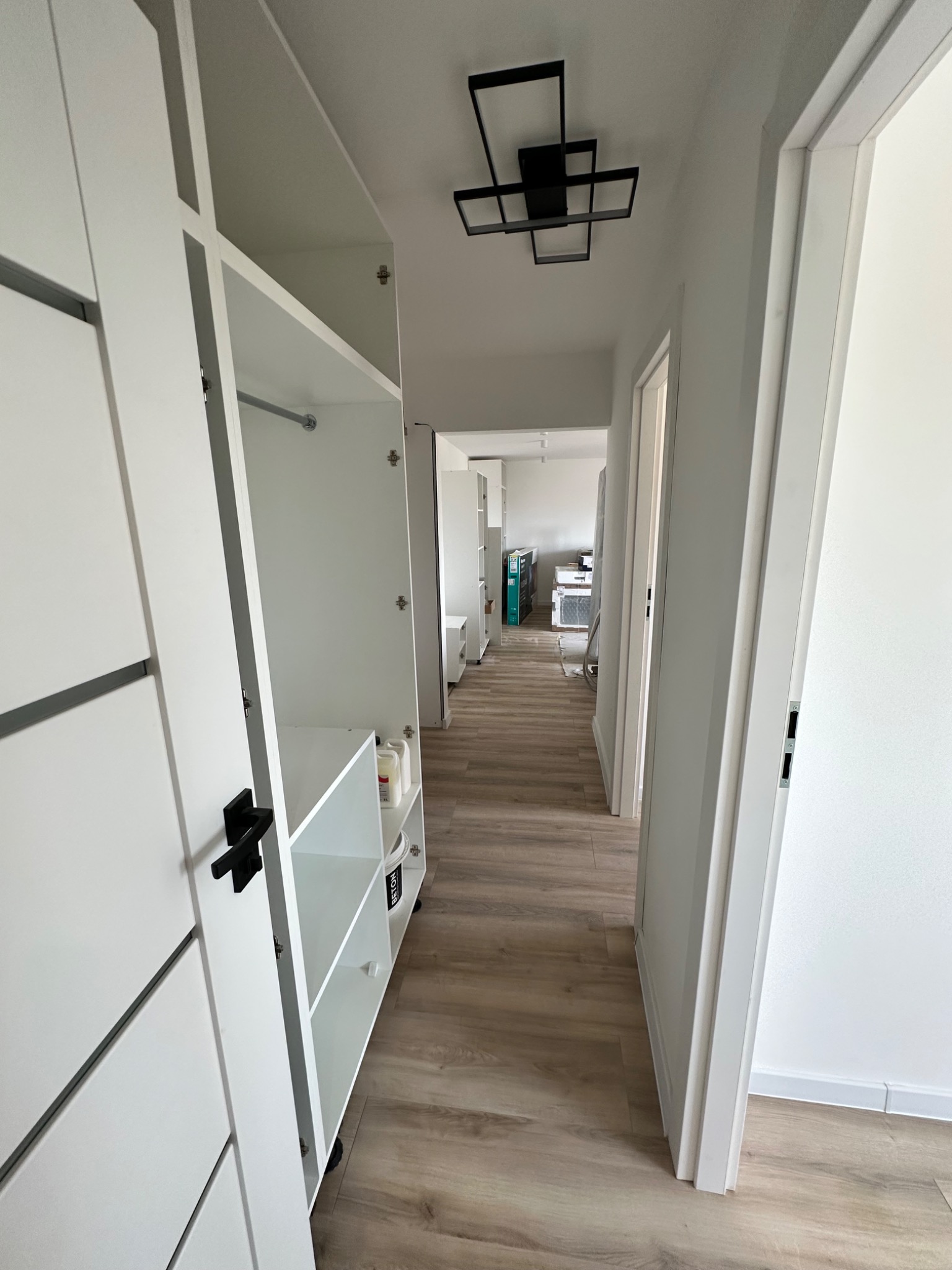 Biała szafa wnękowa z otwartymi drzwiami, widoczne półki i pręt na ubrania, w tle jasne drewniane panele podłogowe i nowoczesna lampa sufitowa.