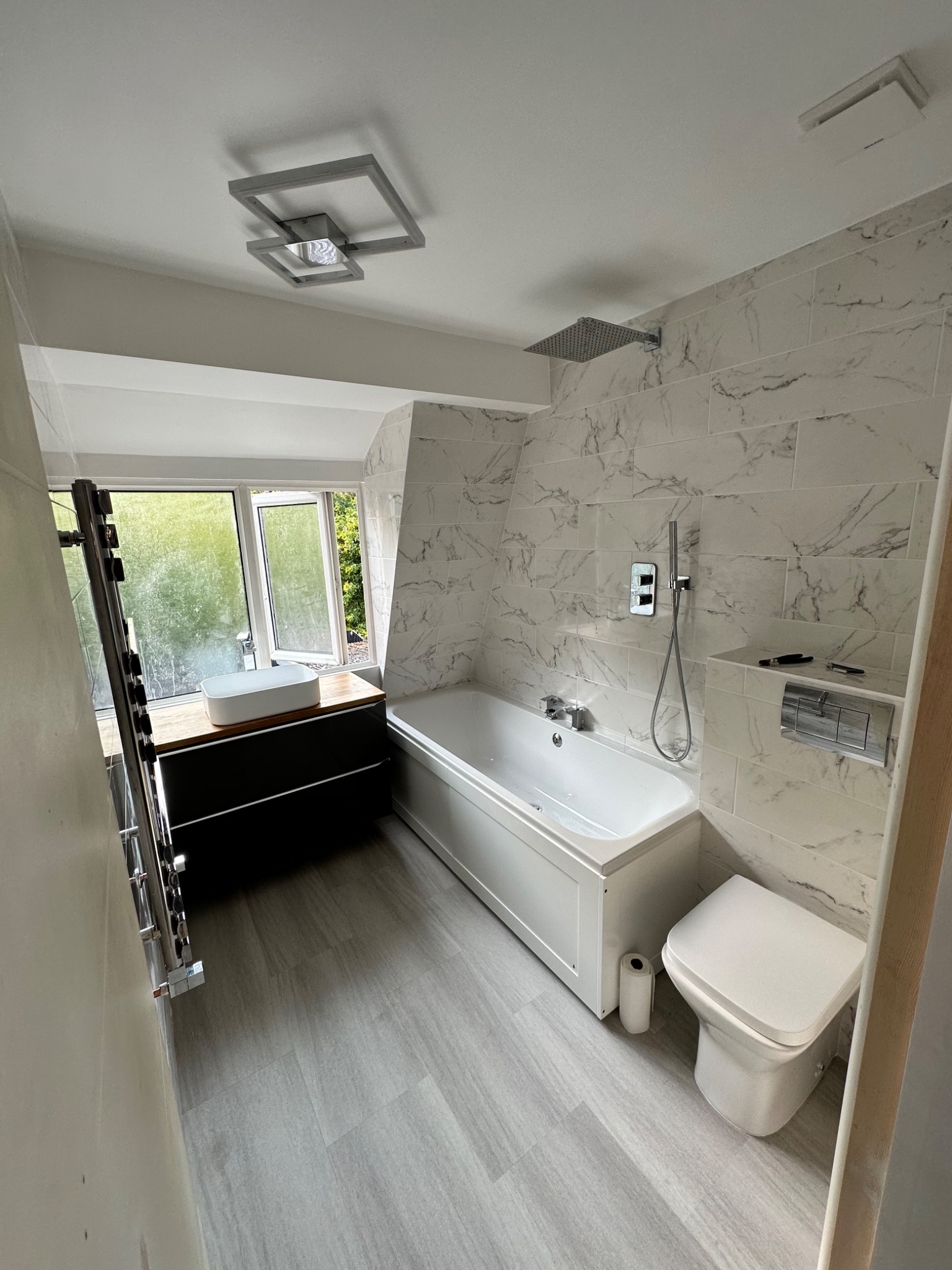 Nowoczesna łazienka z marmurowymi płytkami, białą wanną, umywalką nablatową na ciemnej szafce, grzejnikiem drabinkowym i podłogą imitującą drewno.