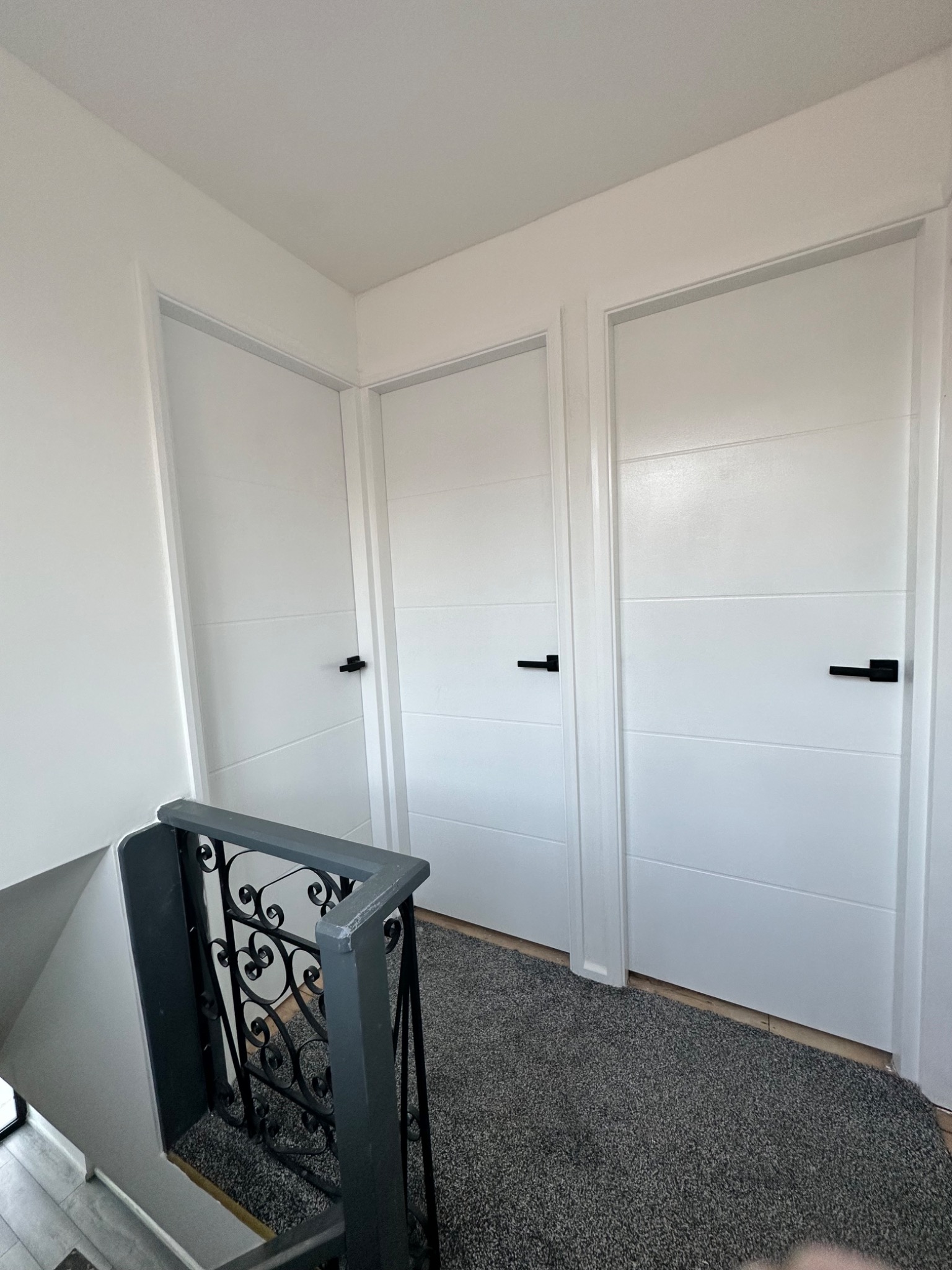 Nowoczesne, białe drzwi wewnętrzne z poziomymi frezowaniami i czarnymi klamkami, widok z góry schodów z ozdobną, metalową balustradą.