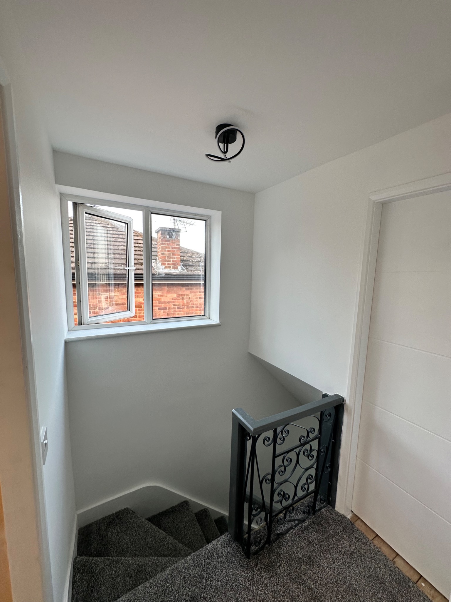 Wnętrze klatki schodowej po remoncie: świeżo pomalowane ściany w odcieniach bieli i szarości, widoczne schody pokryte szarą wykładziną, ozdobna metalowa barierka i okno z widokiem na ceglany komin.