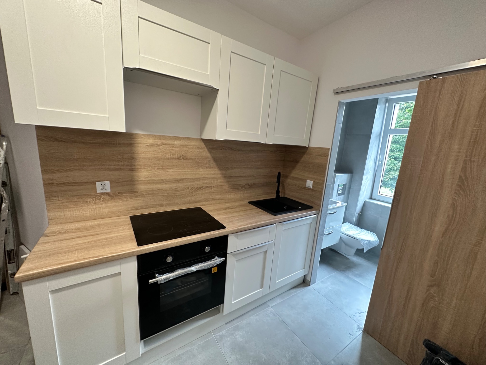 Nowoczesna, jasna kuchnia z drewnianym blatem i czarnym zlewem oraz płytą indukcyjną, widok na częściowo zasłoniętą łazienkę z toaletą w tle.