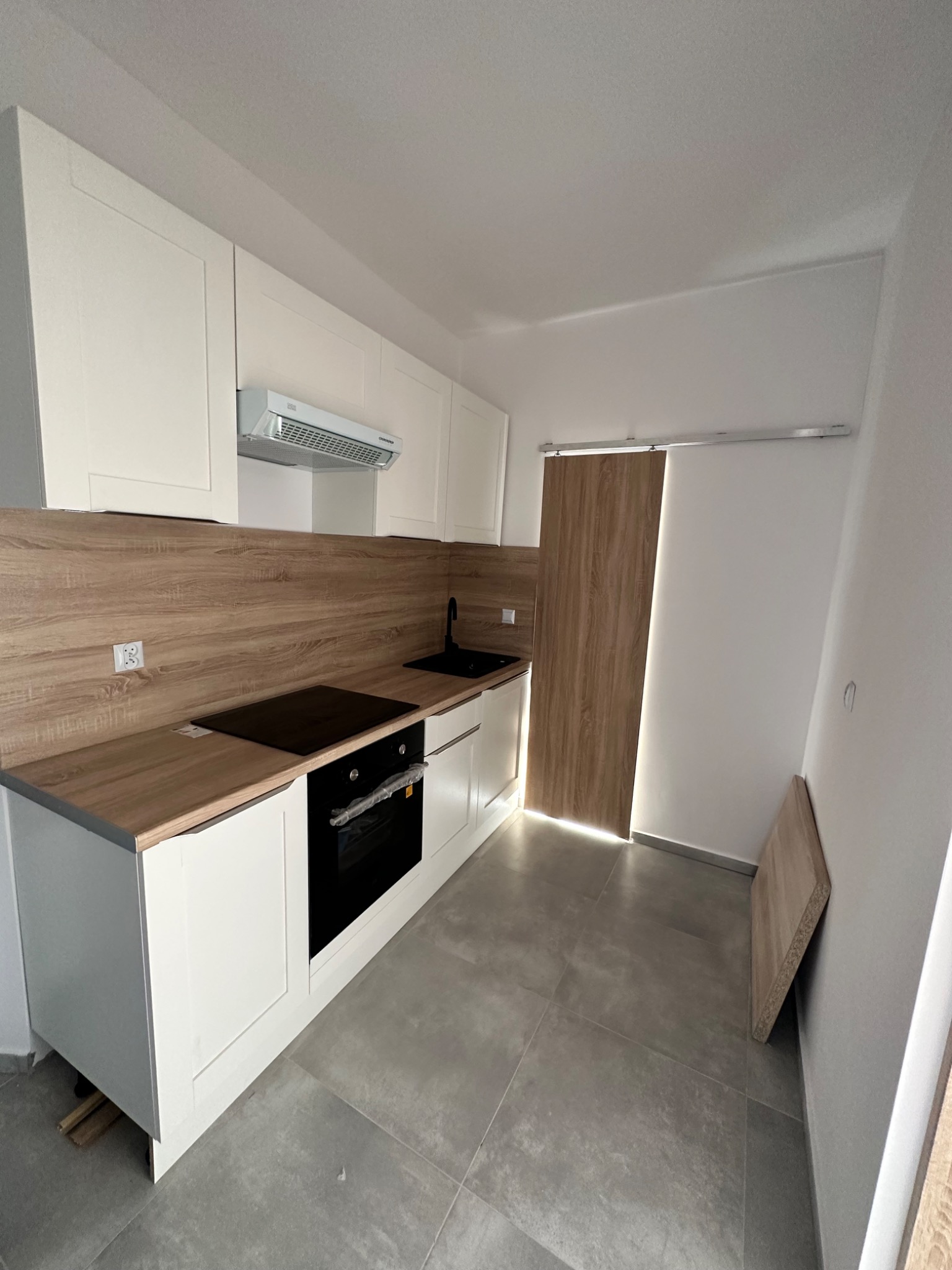 Nowoczesna kuchnia w trakcie wykańczania: białe szafki z drewnianym blatem, czarna płyta indukcyjna i zlew, szare płytki podłogowe, przesuwne drzwi z drewnopodobnym dekorem, element płyty oparty...