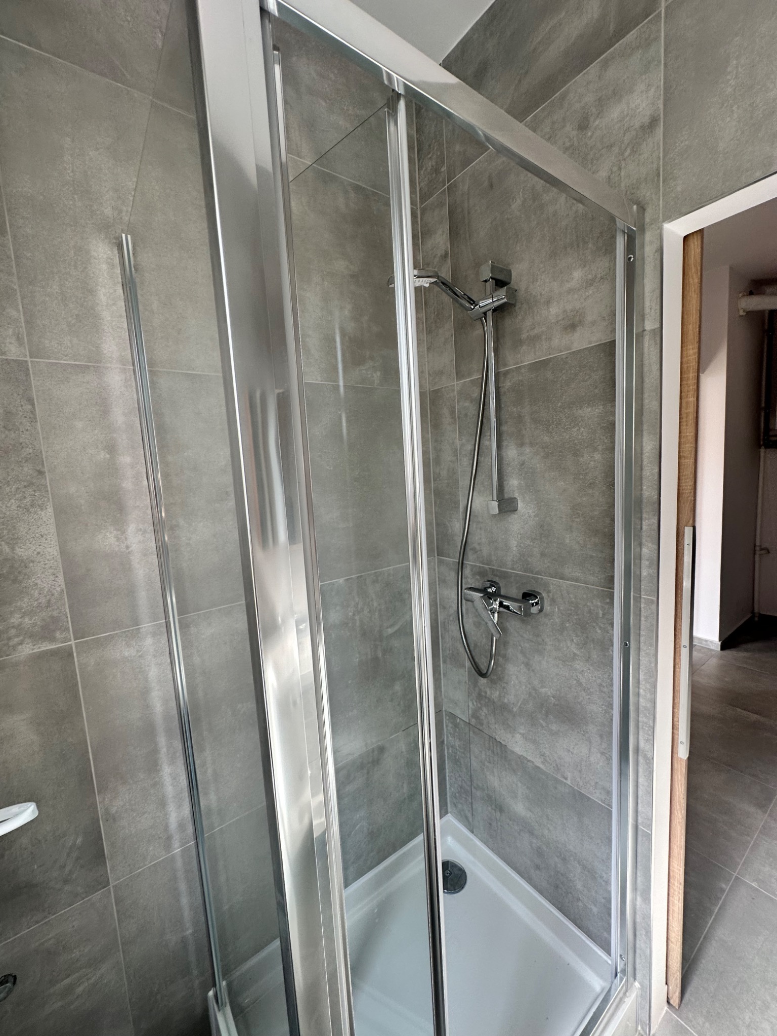 Szklana kabina prysznicowa z chromowanymi profilami i szarymi płytkami w nowoczesnej łazience, widok na biały brodzik i baterię.