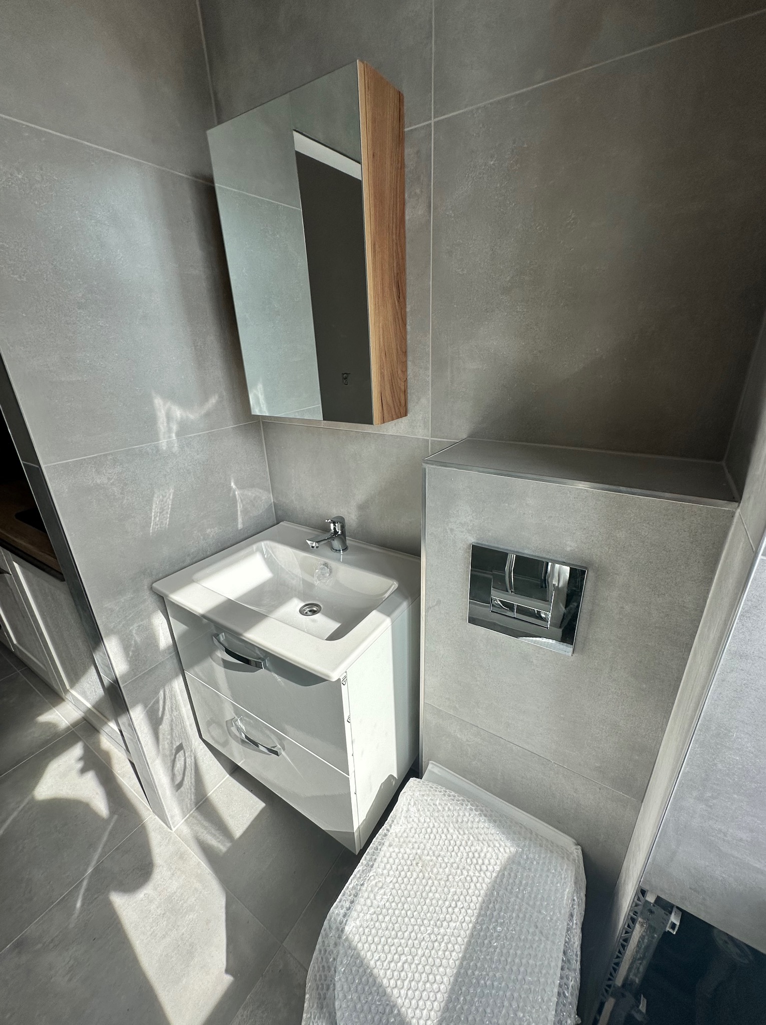Szara łazienka z białą szafką pod umywalką, lustrem z drewnianym akcentem i toaletą zabezpieczoną folią bąbelkową.