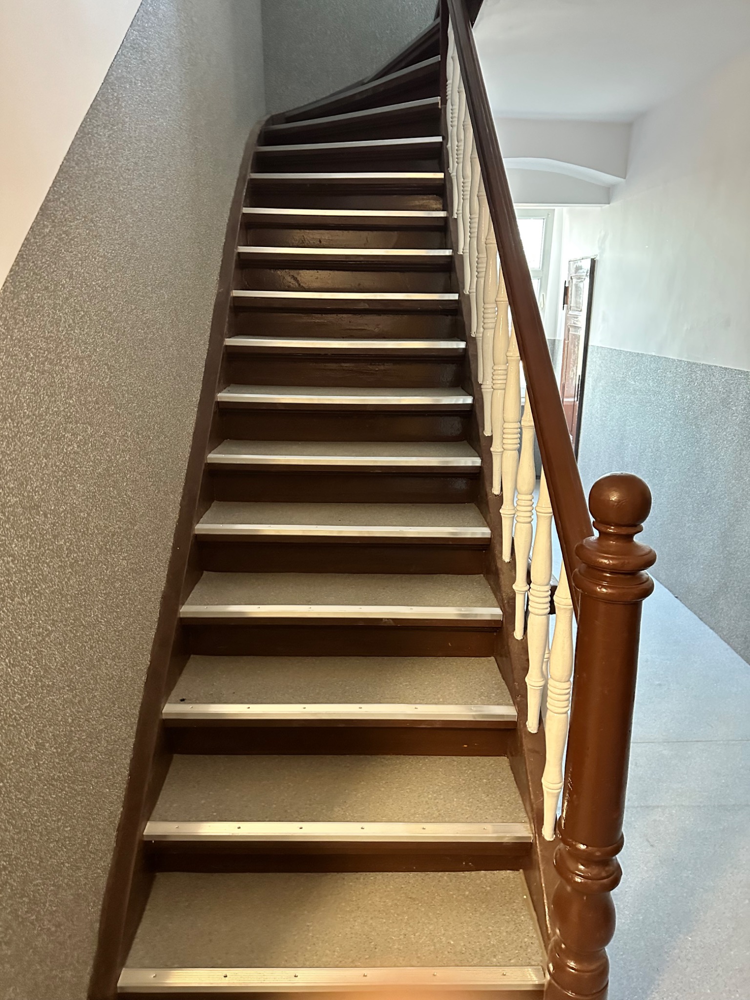 Kręte schody z brązowymi stopniami i białymi balustradami, wykończone aluminiowymi listwami antypoślizgowymi, prowadzące w górę w budynku o ścianach pokrytych szarym tynkiem.