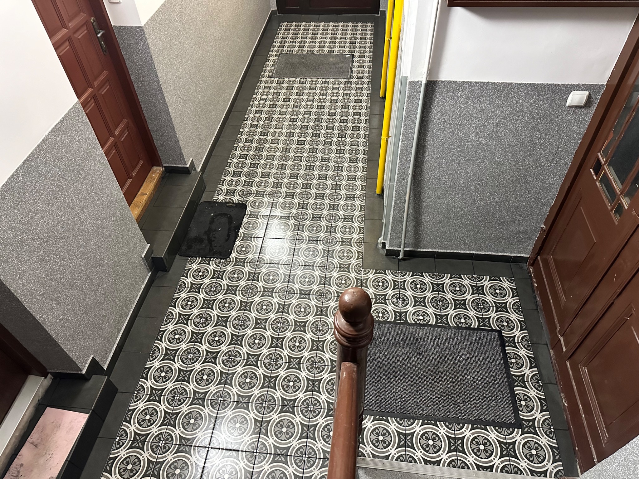 Korytarz po remoncie z nową, wzorzystą podłogą z płytek, świeżo pomalowanymi ścianami z fakturą baranka i brązowymi drzwiami wejściowymi.