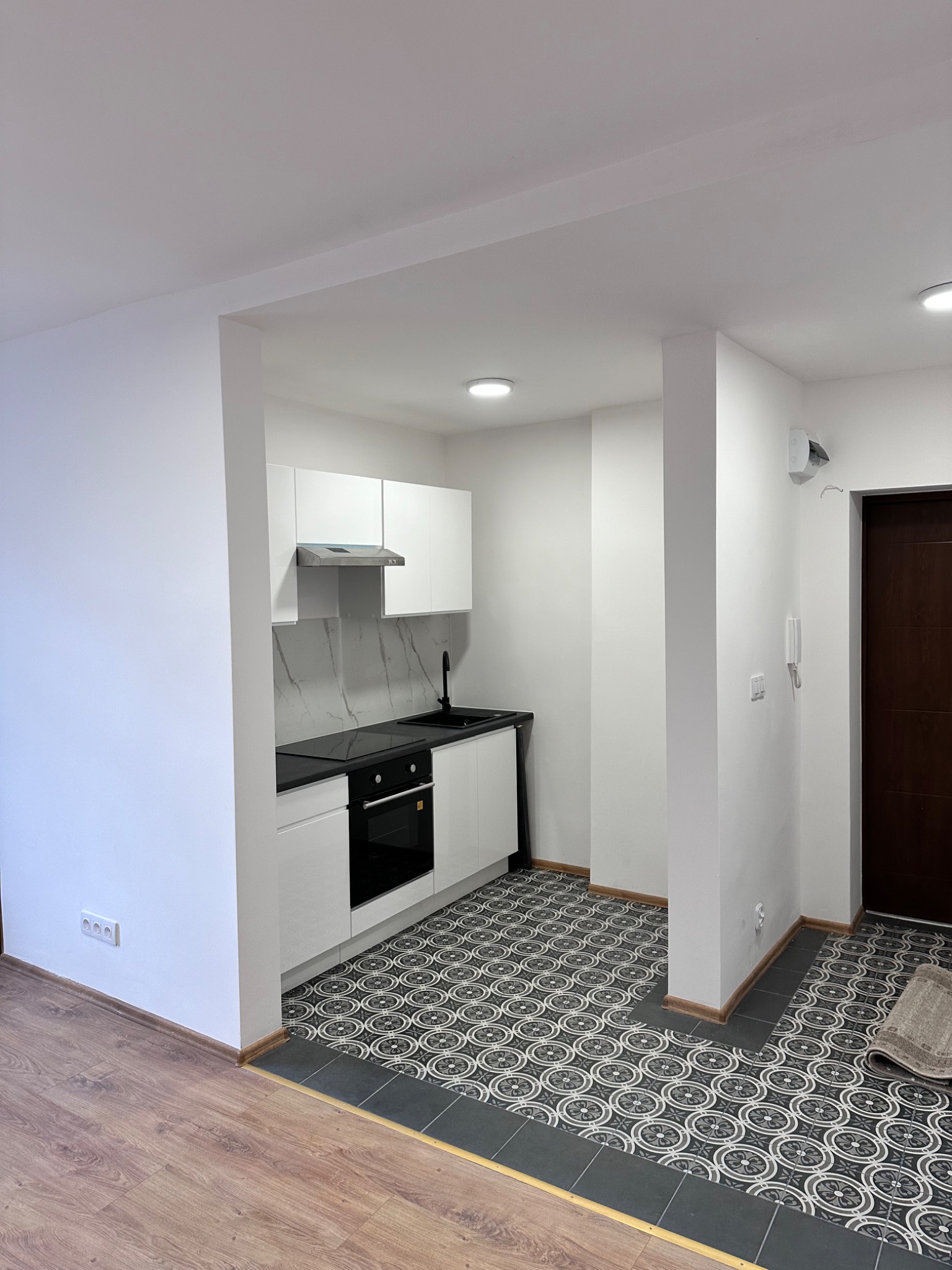 Nowoczesna, minimalistyczna kuchnia z białymi szafkami, czarnym blatem i płytkami podłogowymi w geometryczny wzór, widok z korytarza z drewnianą podłogą.