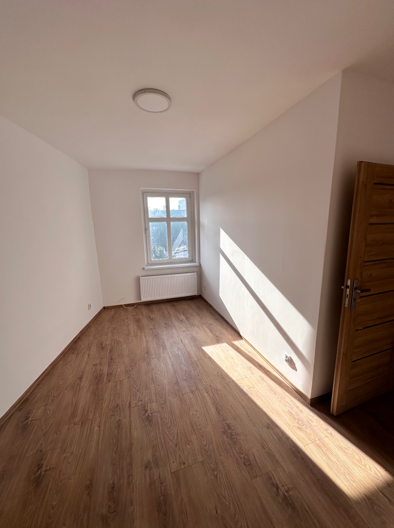 Pusty pokój po remoncie z drewnianą podłogą, świeżo pomalowanymi białymi ścianami i oknem z grzejnikiem, zalany popołudniowym światłem słonecznym wpadającym przez drzwi.