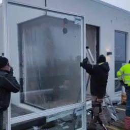 Montaż okna panoramicznego z ramą aluminiową przez dwóch pracowników w odzieży roboczej, widoczne przyssawki do szkła i drabina oparta o ścianę budynku.