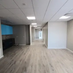 Wnętrze biurowe z panelami podłogowymi imitującymi drewno, sufit podwieszany kasetonowy z wbudowanym oświetleniem LED, ściany w odcieniach szarości, niebieska zabudowa kuchenna w oddali.