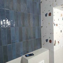 SERHII STARODUBTSEV AWANGARD - Błękitne, pionowe płytki ścienne z połyskiem, kontrastujące z mozaikową ścianą w stylu lastryko, telefon leżący na obudowie stelaża podtynkowego WC.