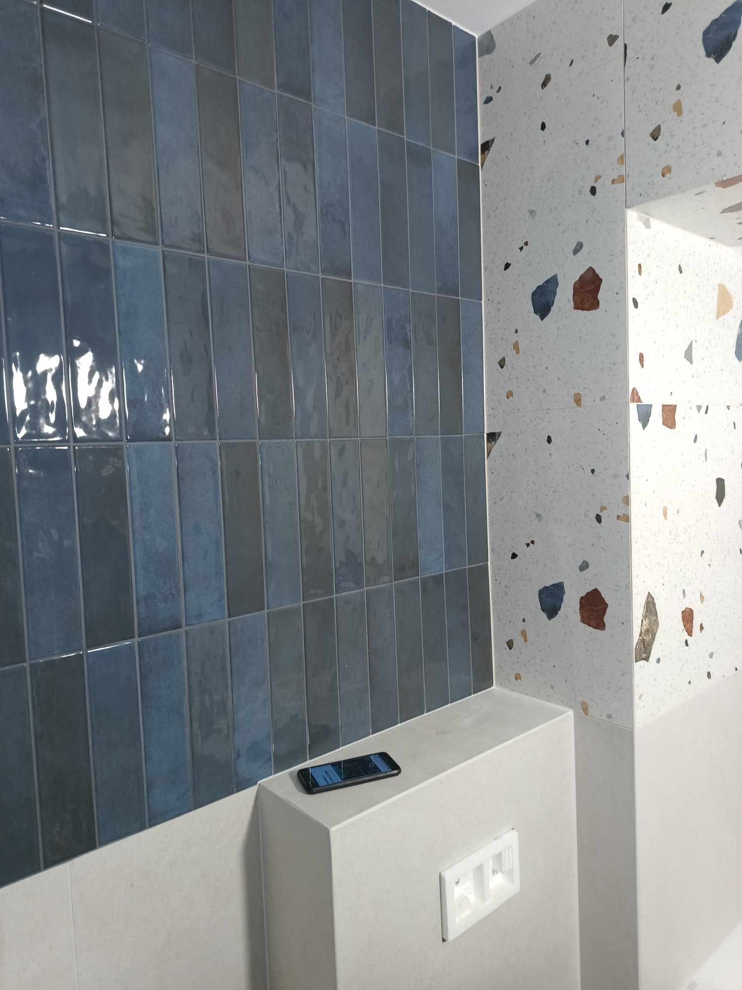 Błękitne, pionowe płytki ścienne z połyskiem, kontrastujące z mozaikową ścianą w stylu lastryko, telefon leżący na obudowie stelaża podtynkowego WC.